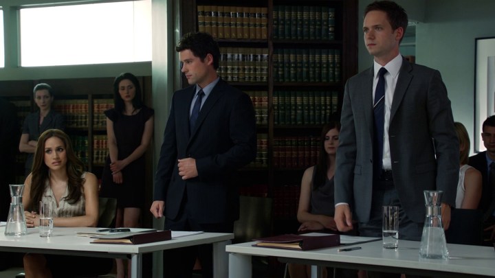 Mike Ross in un finto processo in piedi, Rachel seduta sul lato opposto con un altro avvocato in una scena di Suits.