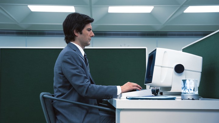 Una vista laterale di Adam Scott seduto alla scrivania con un computer nella serie Severance di Apple TV+.